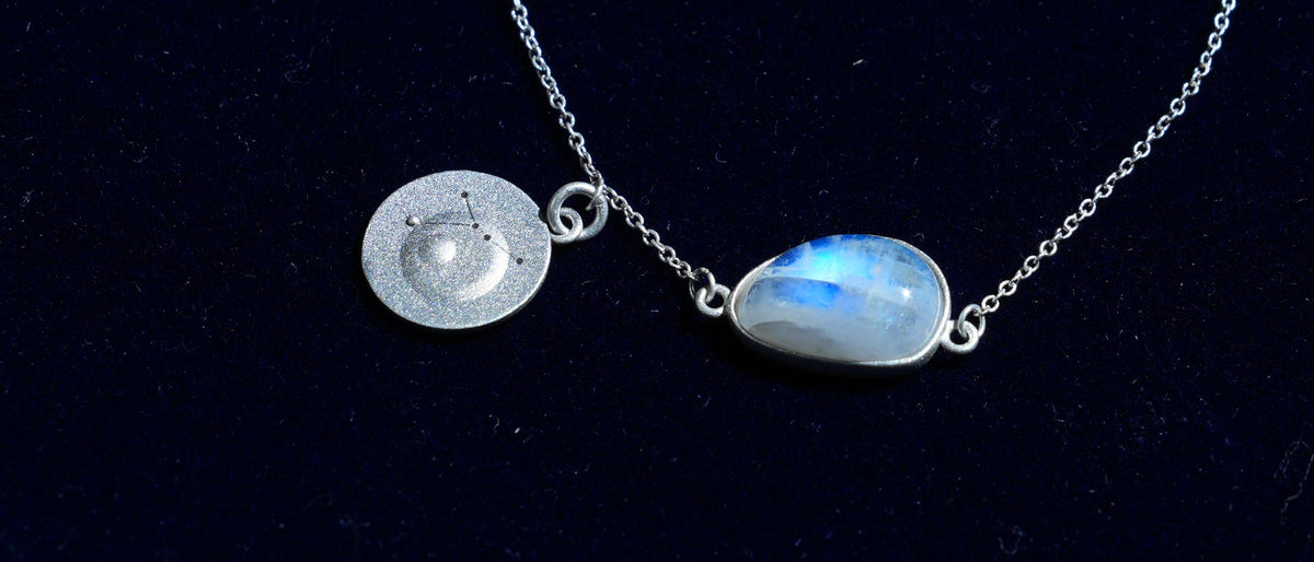 Close-up of the Cancer piece: a natural rainbow moonstone and its constellation coin on navy velvet