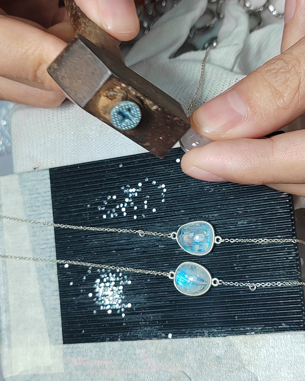 Hands filing a rainbow moonstone setting at the bench, with loose stones and finished bracelets beside