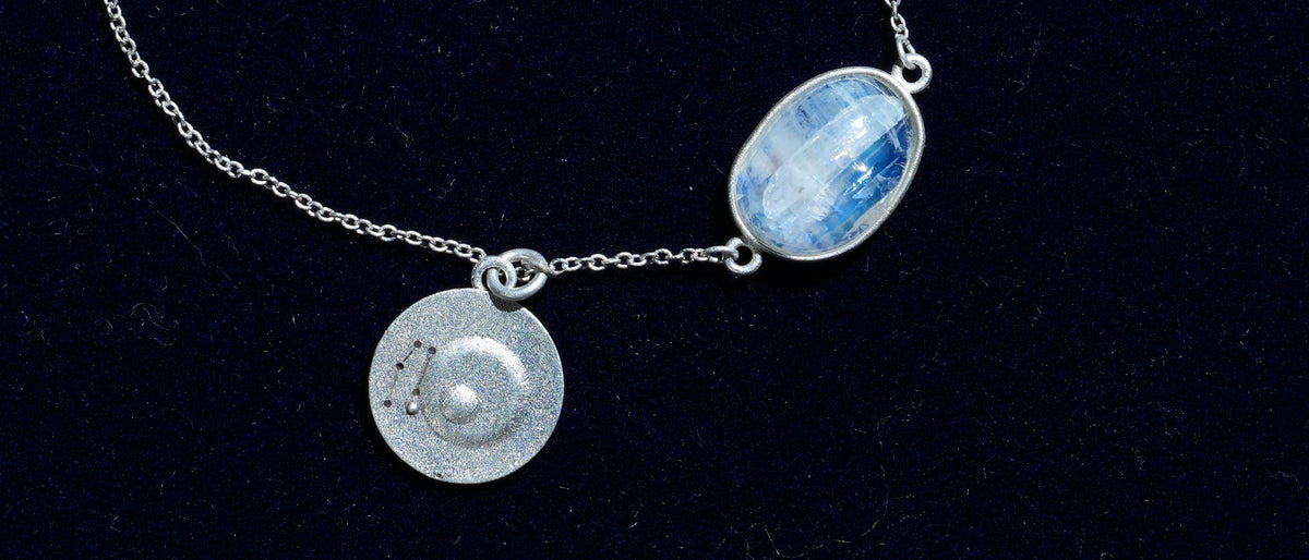Close-up of the Gemini piece: a natural rainbow moonstone and its constellation coin on navy velvet