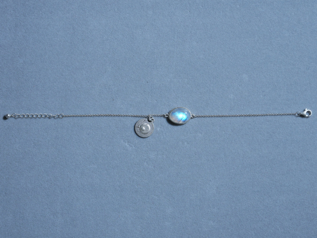 A rainbow moonstone bracelet laid out straight on blue-gray paper, the stone and its coin at the center