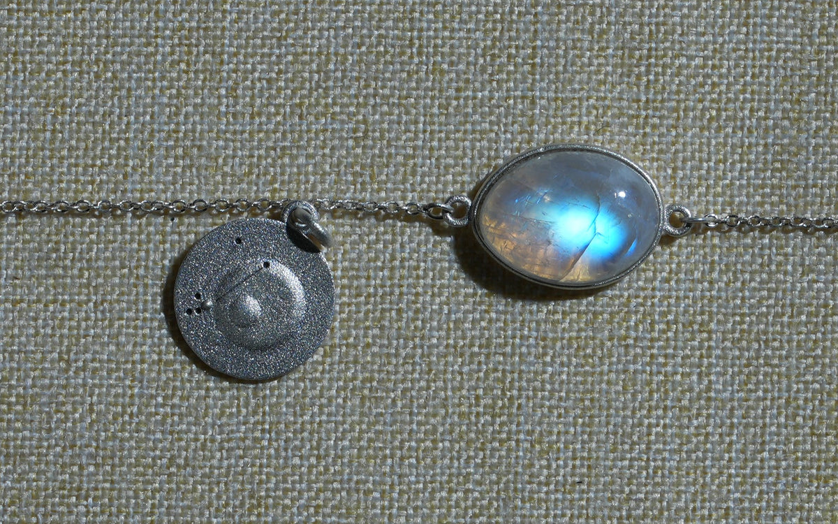 Close-up of an oval rainbow moonstone with a blue flash, beside an engraved silver coin on woven fabric